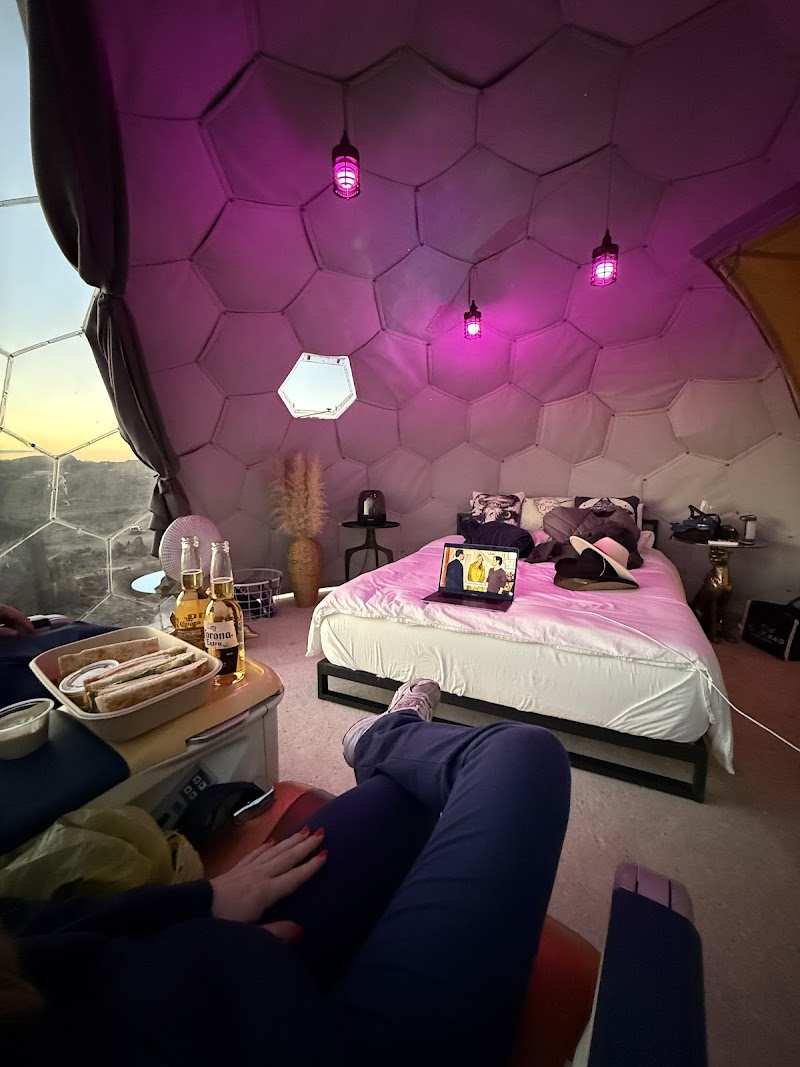 Inside a geodesic dome lodging at Big Bend National Park, a bed with pillows sits under pink ambient lighting and a laptop on the foot of the bed.