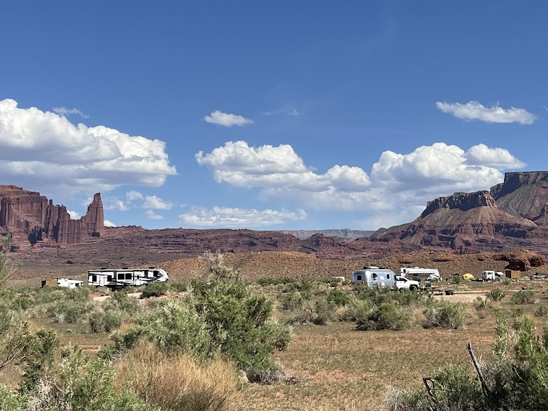 Arches National Park campground with RVs and trailers parked among shrubs, red rock mesas in the distance under a blue sky.