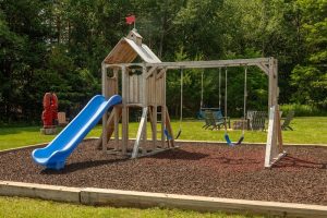 Wooden playground with blue slide and swings on a mulch surface, surrounded by trees in Acadia National Park.