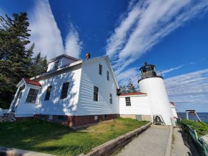 White lighthouse with an attached house sits on a grassy overlook beside the Atlantic in Acadia National Park, under a bright blue sky.