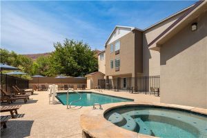 Sunny pool and circular hot tub beside beige hotel buildings at Arches National Park, with lounge chairs and a blue sky.