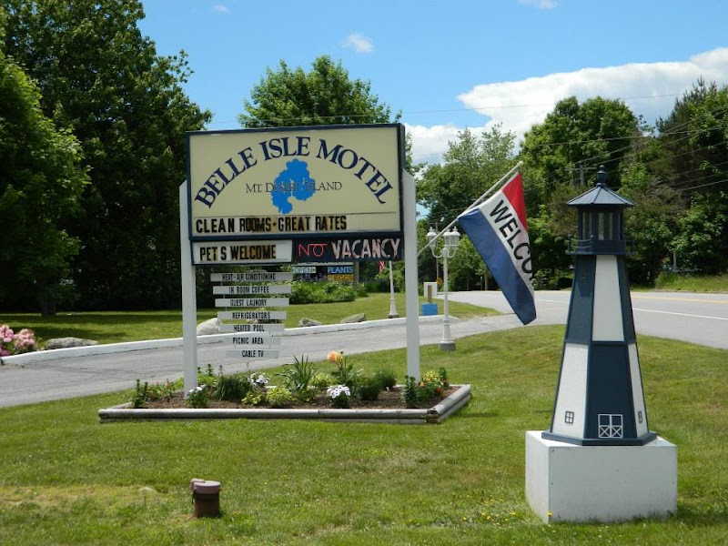 Motel sign with Belle Isle Motel text and map icon, green lawn, flower bed, flag, and a lighthouse sculpture beside a road in Acadia National Park.