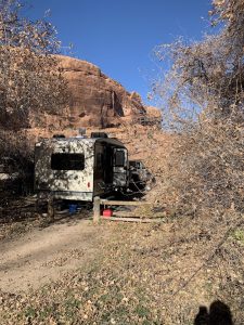 RV trailer beside a wooden fence, with red rock cliffs, blue sky, and leafless shrubs at Arches National Park campground.