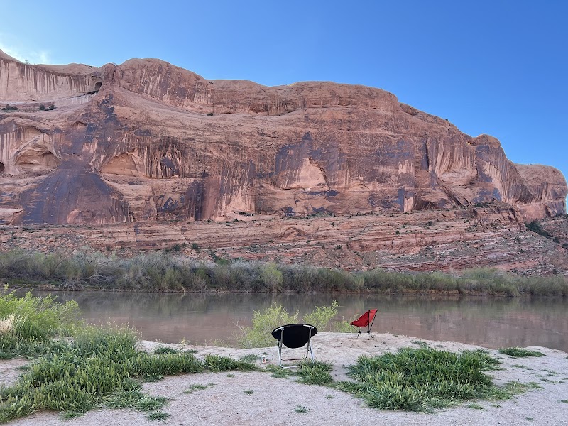 Red sandstone cliffs rise over a calm riverbank with two camping chairs on the shore, Arches National Park.