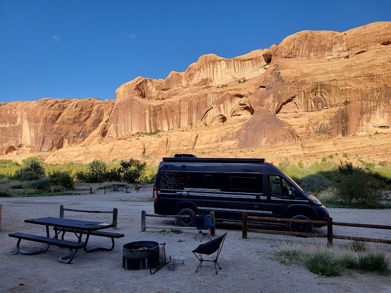 Dark camper van beside a picnic table and campfire ring, with folding chairs, in Arches National Park cliffs.