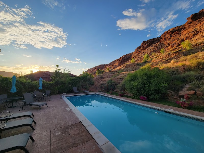 Poolside lounge chairs and blue umbrellas overlook a reflecting pool with red rock cliffs of Arches National Park.