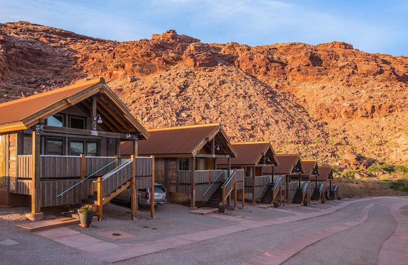 Row of wooden lodge cabins with porches and stairs along a dusty road under orange cliffs at Arches National Park.