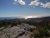 Cracked granite rocks overlook Acadia National Park, with pine shrubs, rolling hills, and a distant ocean coastline under a blue sky.