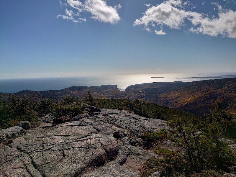 Cracked granite rocks overlook Acadia National Park, with pine shrubs, rolling hills, and a distant ocean coastline under a blue sky.