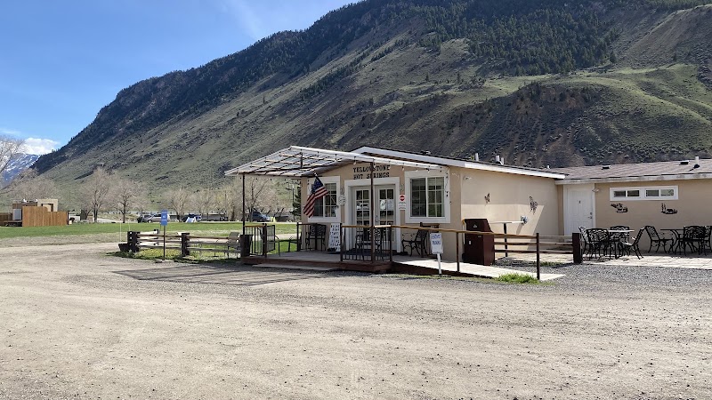 Beige storefront with a porch and outdoor seating, flag at the entrance, on a gravel lot with a green hillside in Yellowstone National Park.