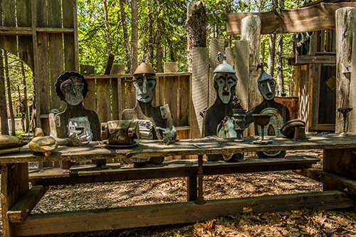 Carved wooden figures and pottery line a rustic display in a gift shop at Acadia National Park, shaded by trees.