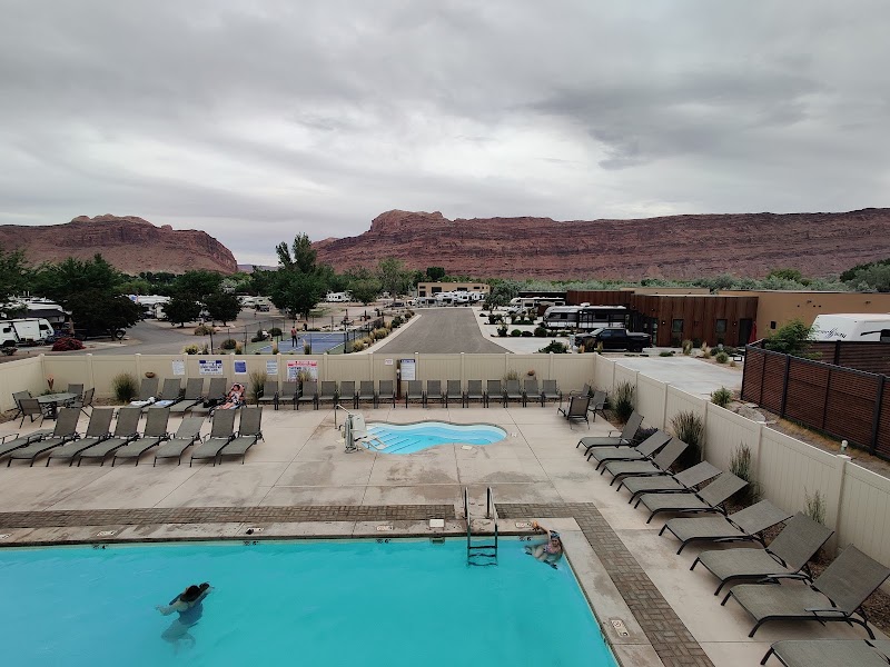 Poolside with lounge chairs around a fenced courtyard, as rugged red rock cliffs of Arches National Park loom beyond RVs and buildings.