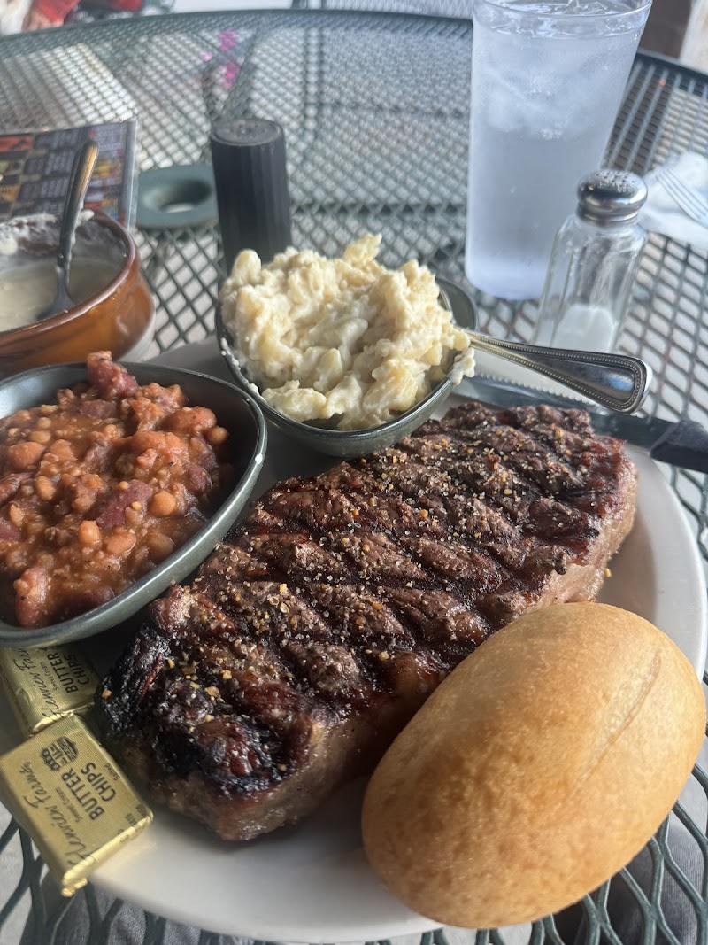 Grilled steak with seasoning, mashed potatoes, beans, a butter packet, and a roll on a plate at Badlands National Park outdoor patio.