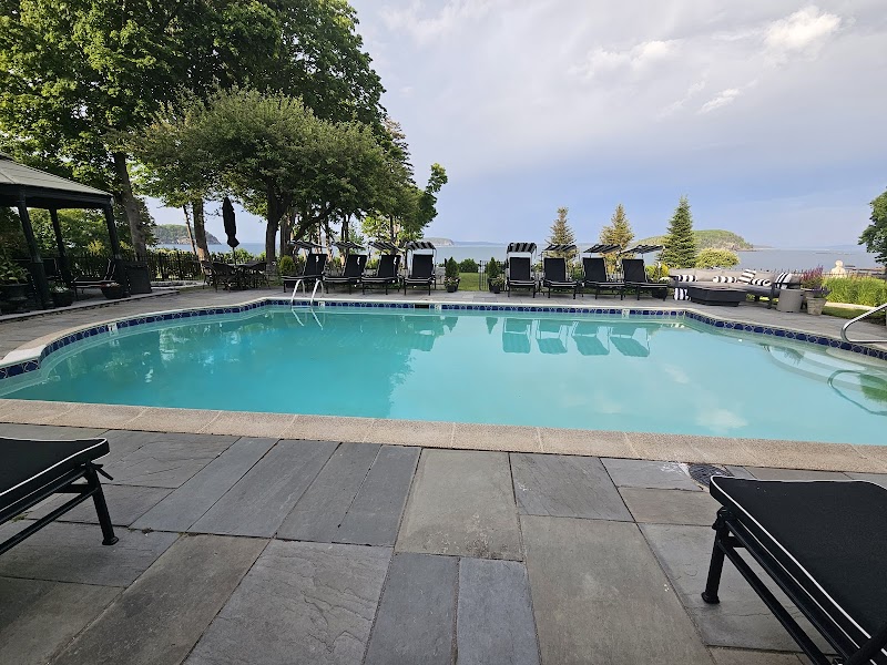 Calm blue pool edged by gray stone patio, rows of lounge chairs, trees and a coastal view at Acadia National Park.