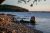Balance Rock stands along the rocky shoreline at Acadia National Park, with calm water and boats in the distance.