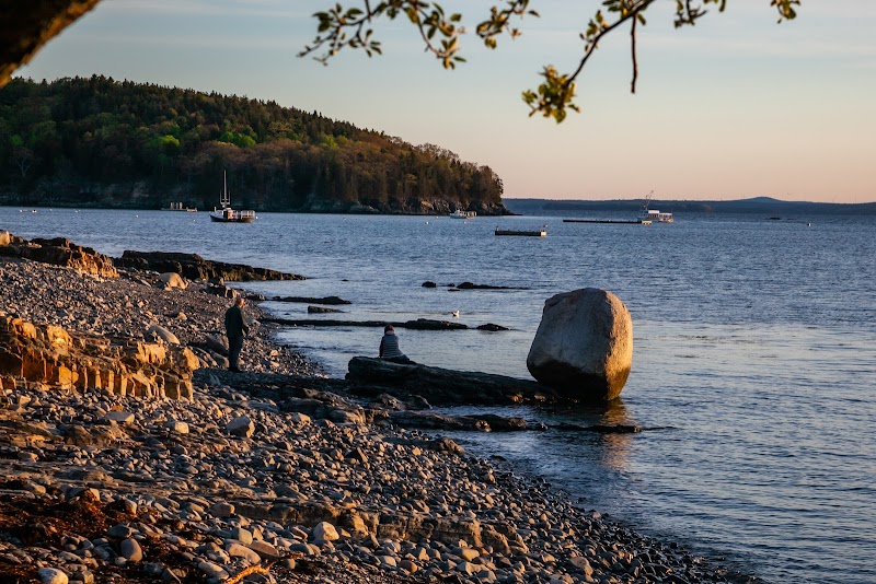 Pebbly shoreline of Acadia National Park with a large balanced rock by the water, a person nearby, boats on the bay at sunset.