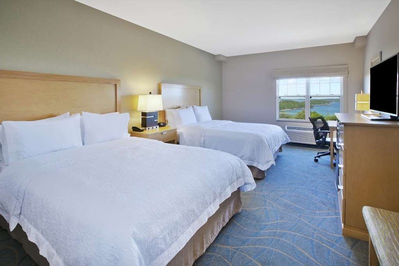 Two queen beds with white linens in a bright hotel room, desk by the window overlooking water in Acadia National Park.