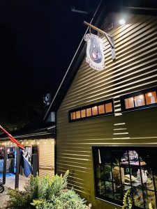 Night exterior of a wood-paneled tavern with a hanging sign featuring a dog and pony, warm windows, string lights, and shrubs in Acadia National Park.