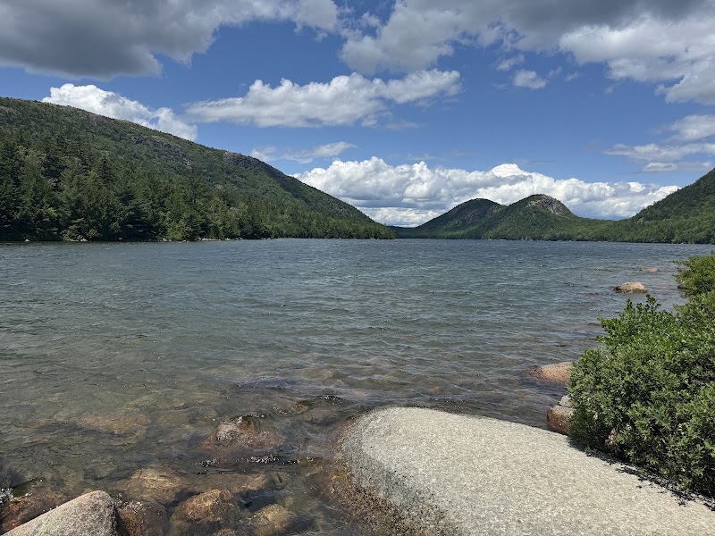 Jordan Pond shoreline at Acadia National Park with clear water, granite shore, and pine-covered hills under a blue sky.