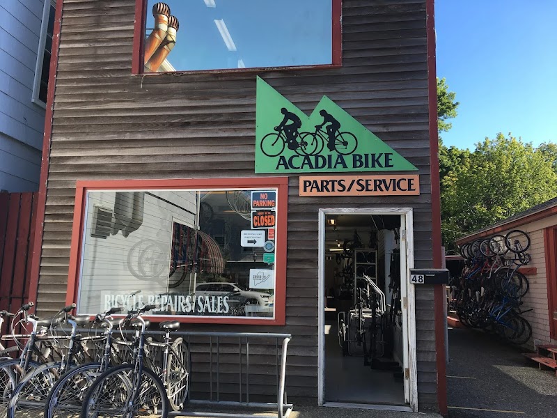 Wooden storefront at Acadia National Park with a green cyclist sign, bikes lined outside, and a partly open door showing shop interior.