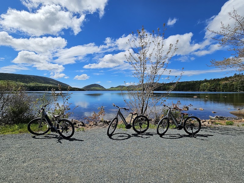 Three bikes lined up on a gravel path beside a calm lake in Acadia National Park, under blue sky and fluffy clouds.