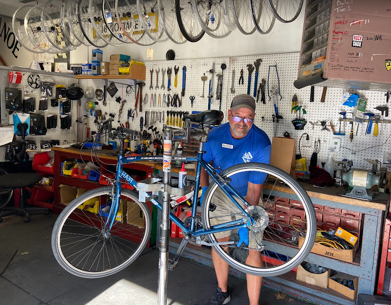 Inside a bike repair shop in Acadia National Park, a man in a blue shirt holds a blue road bike on a stand amid pegboard walls full of wrenches.