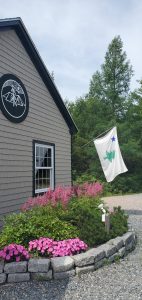 Campground building with circular emblem, pink flowers along a stone border, and a fluttering tree-flag at Acadia National Park.