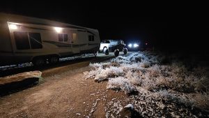 Night at Arches National Park campground shows a large RV and white pickup with headlights on a dirt road beside frosted shrubs.