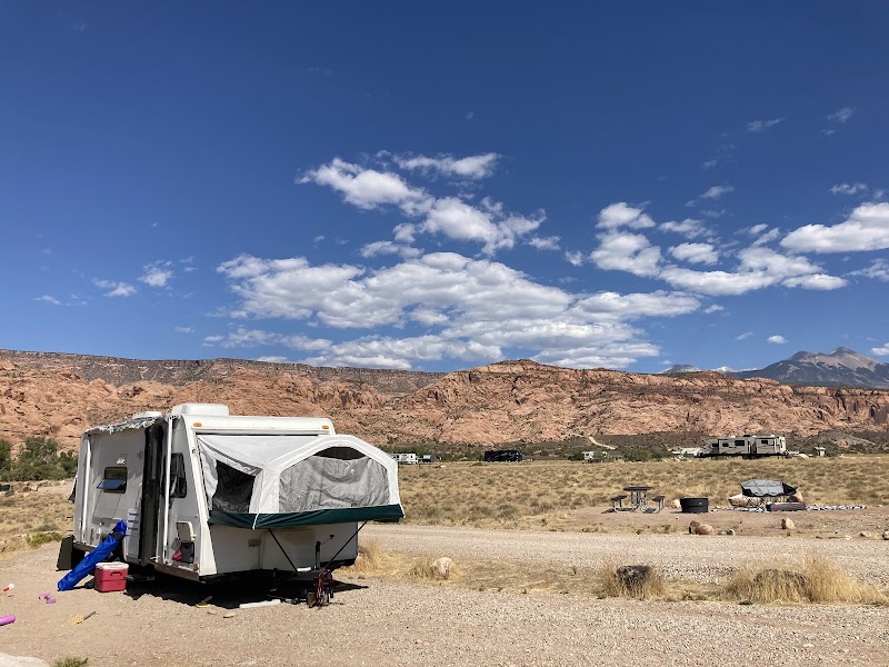 Arches National Park campground with a white trailer and pop-out tent beside red rock mesas under a bright blue sky.