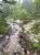 North Bubble Trailhead in Acadia National Park shows a rocky, muddy path with scattered stones, mossy ground, and lush green trees.