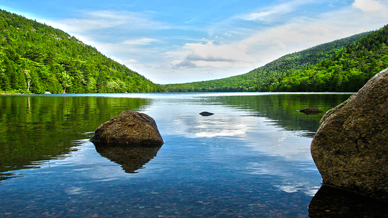 Calm lake at Acadia National Park bordered by forested hills, large rocks in clear blue water under a partly cloudy sky.