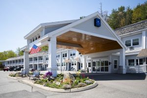 White lodge-style motel with a wooden porte-cochere, flag, benches, and a landscaped circular garden at Acadia National Park.