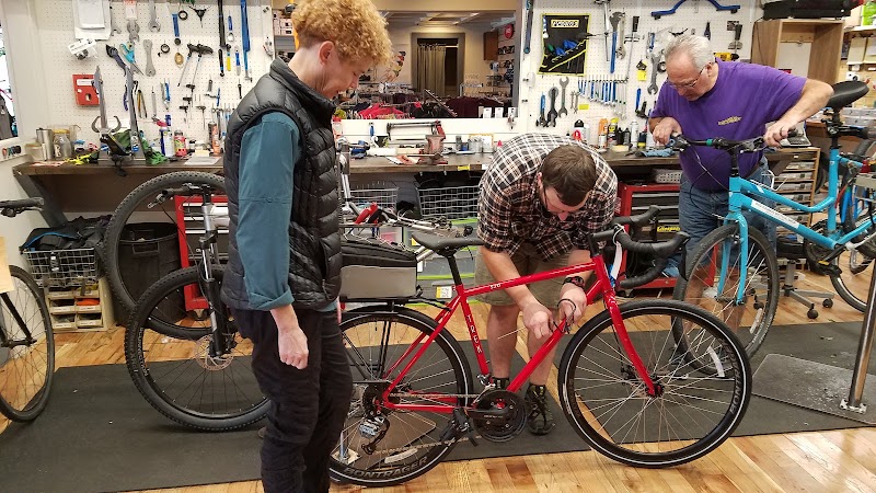 In a busy bike repair shop, three people fix a red road bicycle amid tools and workbenches at Acadia National Park.
