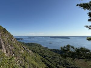 Cliffside overlook in Acadia National Park with a rocky, tree-covered slope on the left, calm blue sea and small islands ahead.
