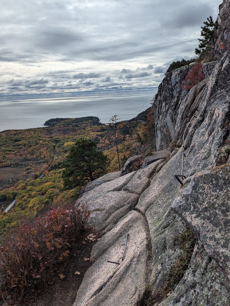 Steep granite trail with metal rungs climbs along a cliff at Acadia National Park, overlooking colorful autumn forest and the distant sea.