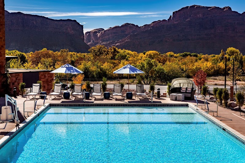 Crystal blue swimming pool with lounge chairs and umbrellas, desert trees and red rock cliffs of Arches National Park in the distance.
