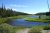 Meandering blue river through grassy green meadows bordered by evergreen forest under a bright blue sky in Yellowstone National Park.
