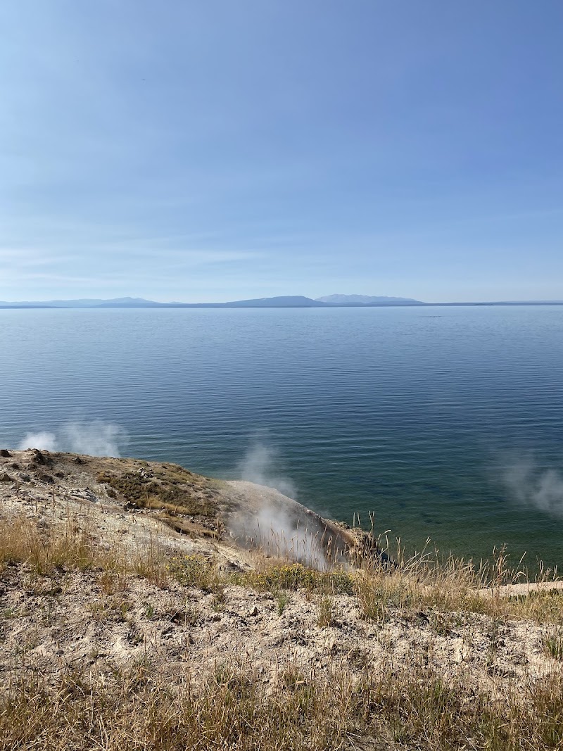 Calm blue lake with distant mountain range, steam rising from rocky shore and dry grasses at Yellowstone National Park.