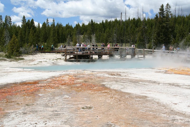 Crowd on a wooden boardwalk over a steaming blue hot spring with orange mineral deposits in Yellowstone National Park.