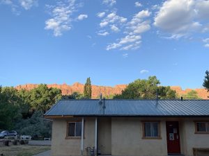 Beige, single-story building with a gray metal roof sits among trees as red rock cliffs rise behind in Arches National Park.