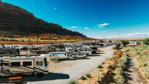 Rows of RVs and campers line a dusty lot under a blue sky with red rock cliffs near Arches National Park.