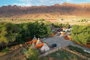 Aerial view of a campground with teepees, trees, and vehicles, backed by red rock cliffs in Arches National Park.