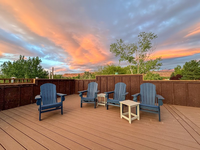 Sunset skies over a wooden deck with five blue Adirondack chairs and small tables at Arches National Park.