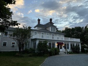 Lodging building with a wraparound porch and dormer windows in Acadia National Park at dusk, surrounded by trees.