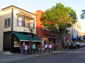 Sunlit street in Acadia National Park with colorful storefronts, a green awning, and people gathered outside.