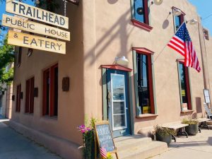 Tan adobe building with blue door, wooden Trailhead signs, American flag, window benches and a chalkboard menu outside in Arches National Park.