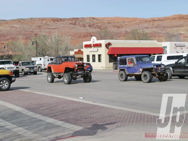 Parking lot with orange and blue off-road jeeps in front of a diner with a red awning, Arches National Park desert cliffs in the background.