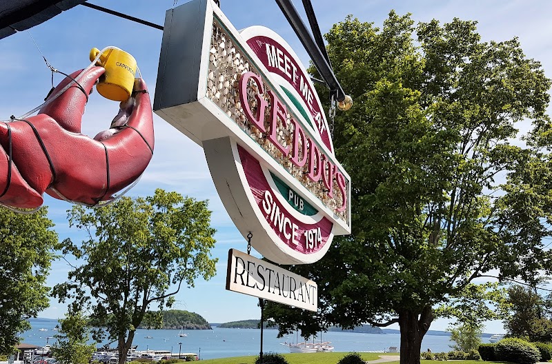 Bright neon restaurant sign with a giant lobster claw hanging above the entrance, overlooking trees and a calm harbor in Acadia National Park.