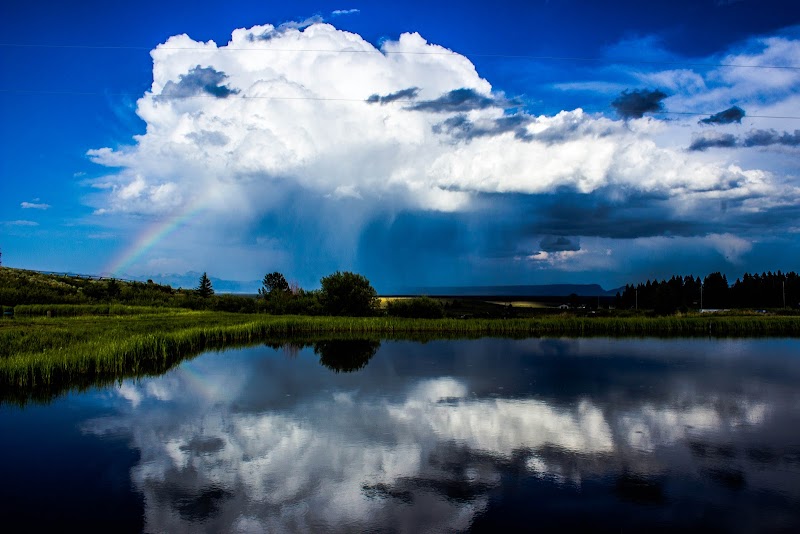 Stormy sky above a calm lake at Yellowstone National Park, rainbow arch, green meadow shoreline, and cloud reflections.