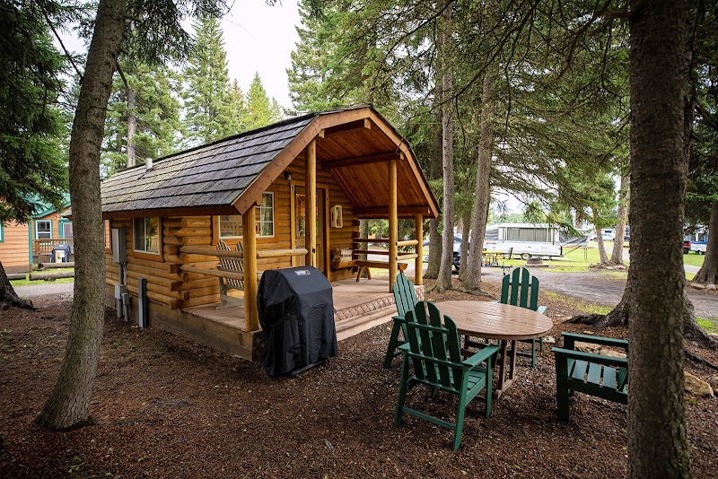 Cozy log cabin with a covered porch, grill, and a round table with green chairs beside pines in Yellowstone National Park.